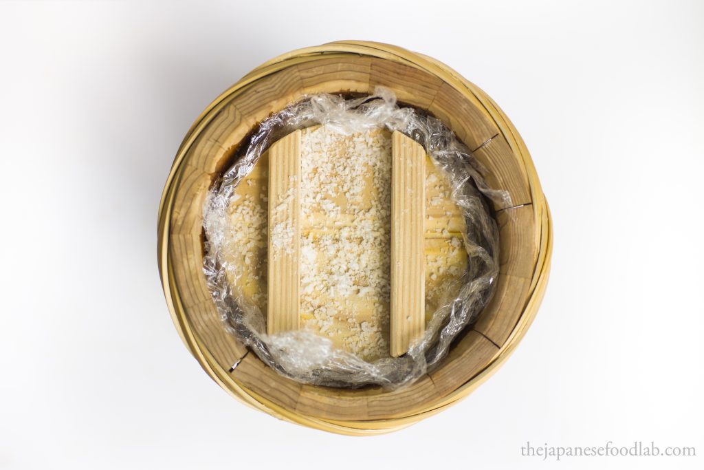Miso covered with clingwrap and  wooden lid with salt fermenting in a wooden barrel.